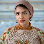 Portrait of a woman in a colorful embroidered outfit, wearing a headscarf and hoop earrings, looking directly at the camera.